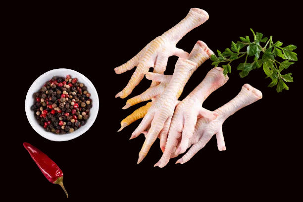 Raw chicken feet on a piece of paper, surrounded by various seasonings and vegetables on a grey surface.