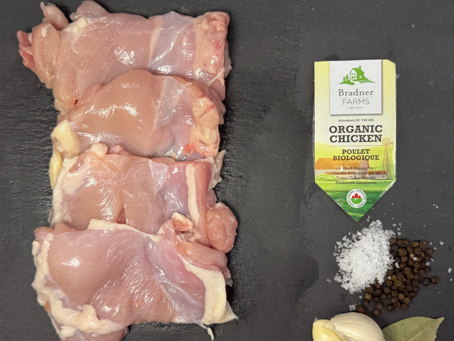 Four raw chicken thighs arranged on a dark slate surface, with a package of Dr. Brands Organic Paprika Seasoning, coarse salt, black peppercorns, a garlic bulb, and a bay leaf below.