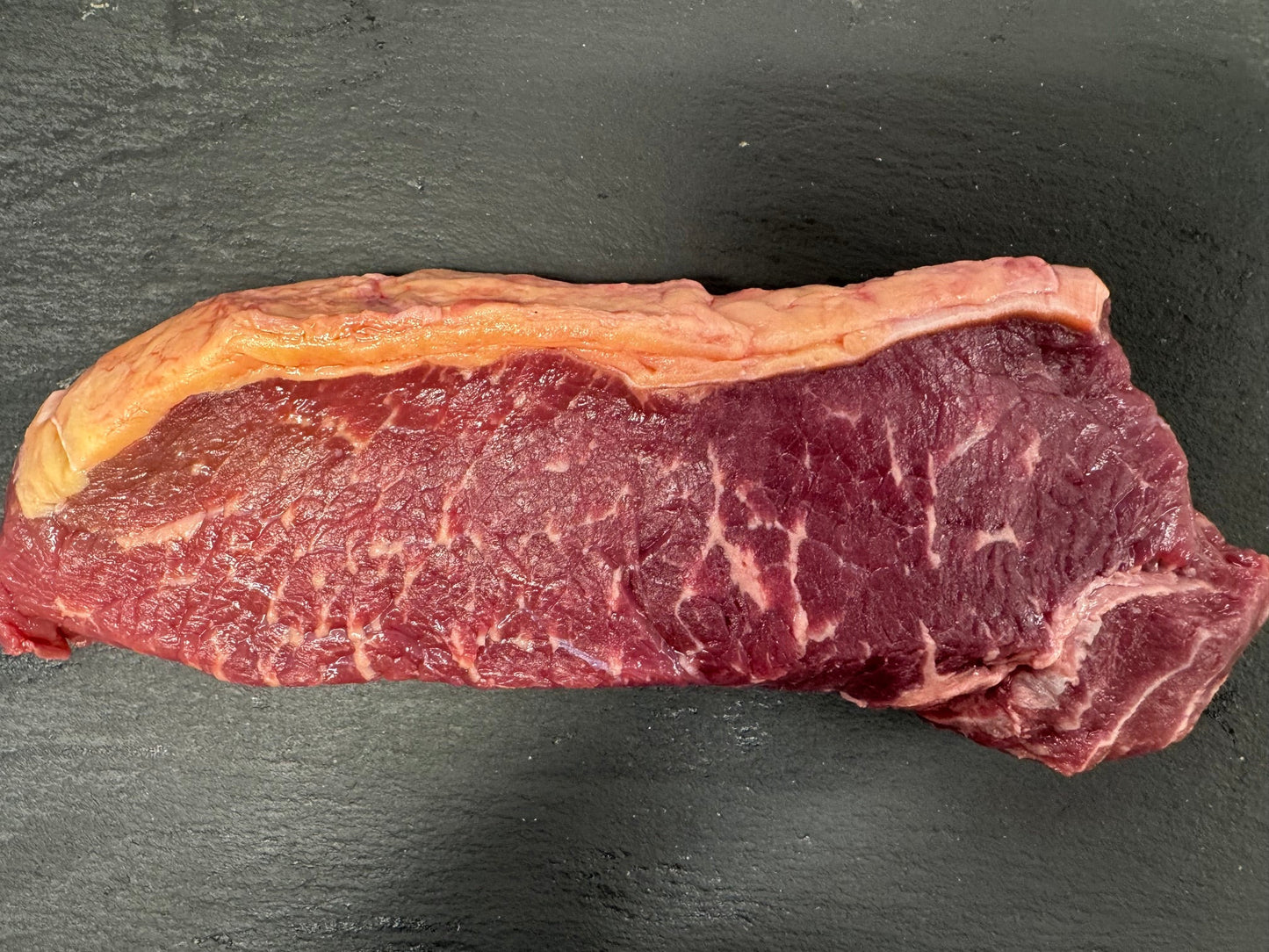 A raw cut of beef steak with marbling and a thick layer of fat on one side, placed on a dark textured surface.