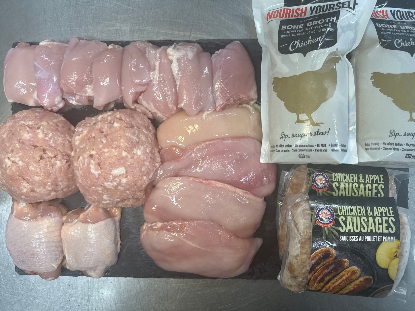 Raw chicken parts and two packaged food items arranged on a dark tray against a stainless steel background. The food includes several raw chicken thighs, two raw chicken breasts, two raw chicken drumsticks, two raw chicken legs, two raw ground meat balls, a package of chicken and apple sausages, and two white packages of chicken wings.