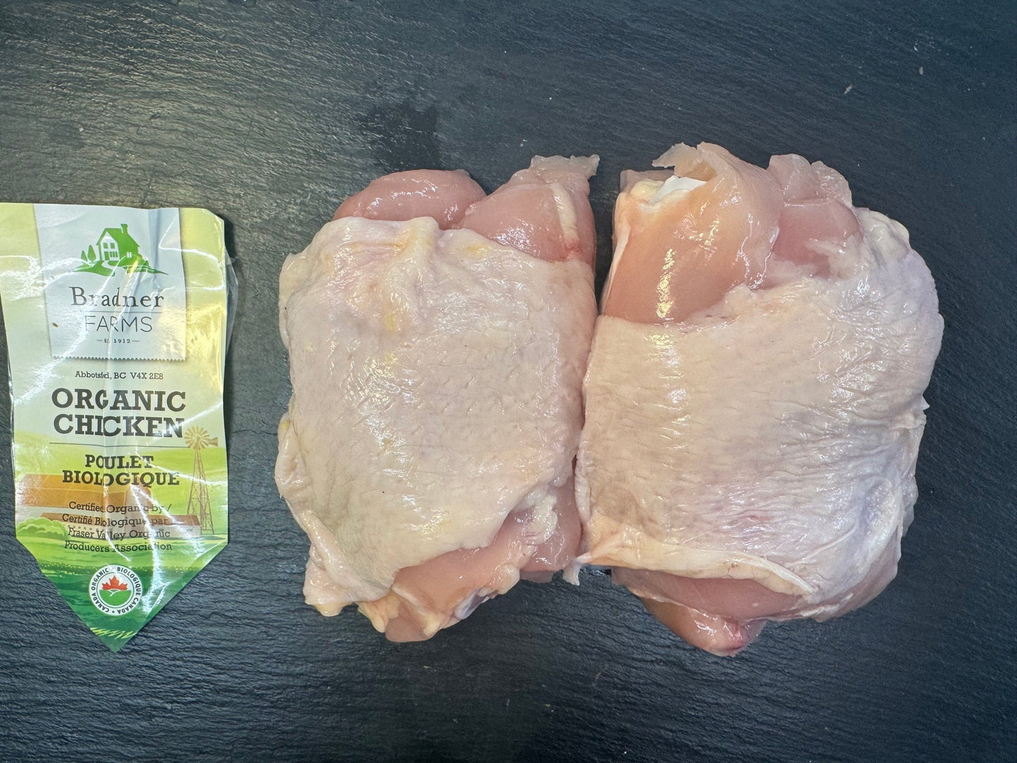 Two raw chicken thighs placed on a dark slate surface with a package of Product of Canada Organic Chicken in the background.
