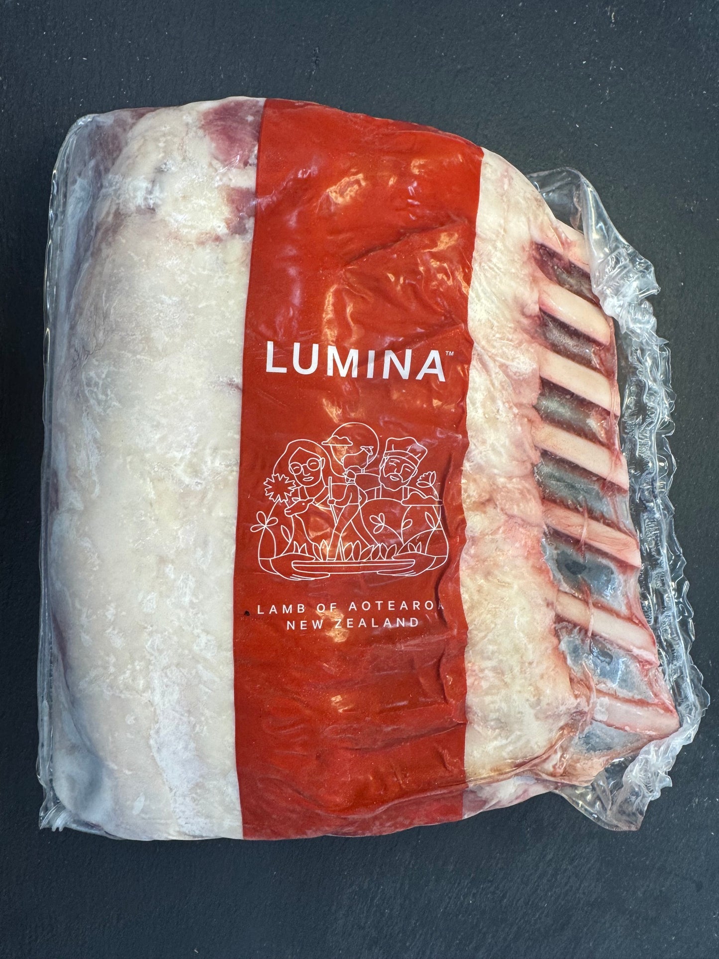 A vacuum-sealed package of Lumina Lamb from New Zealand meat, showing the white marbled fat and red meat layers with visible bones on the side, set against a dark slate background.