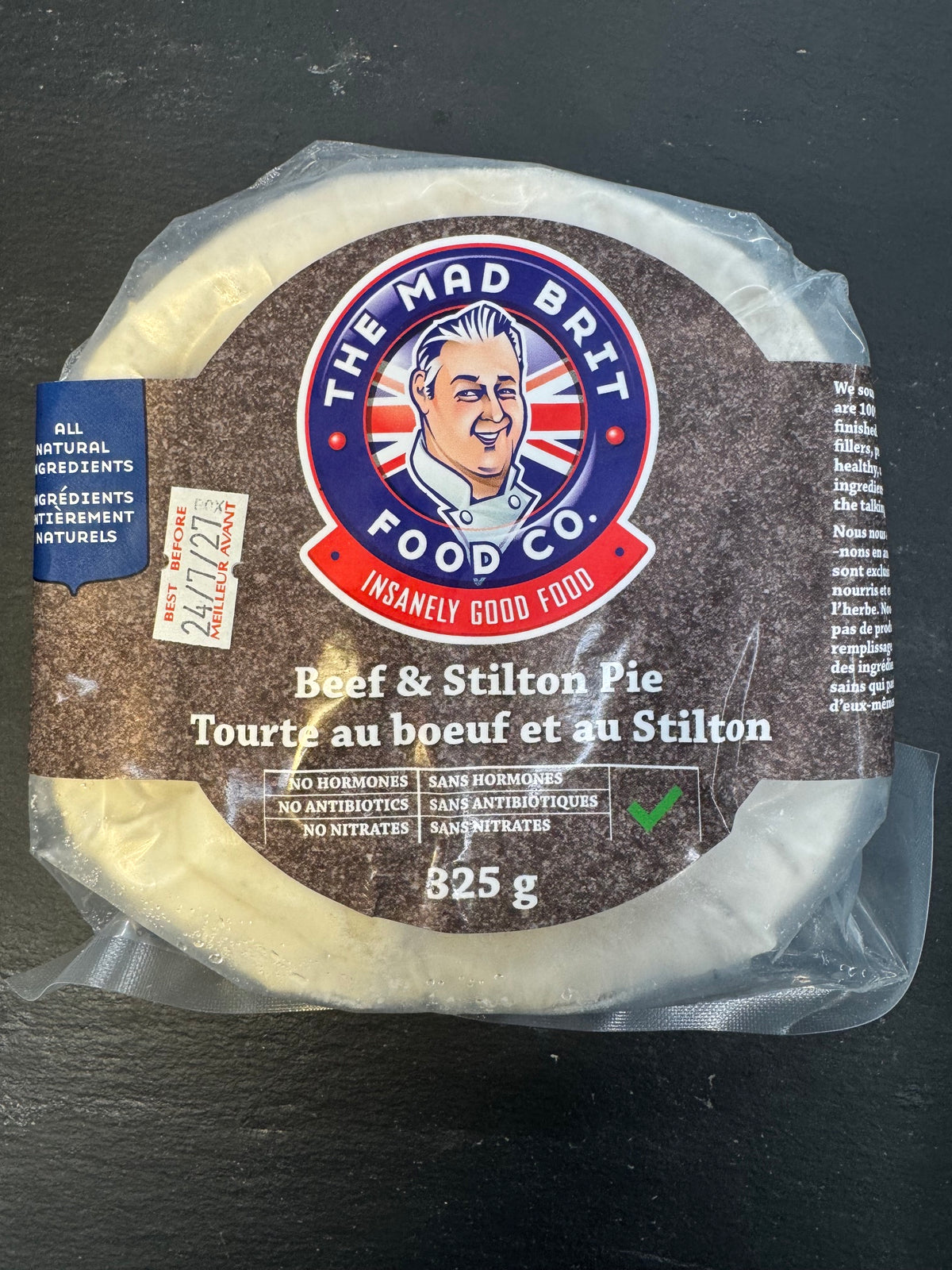 A packaged round cheese product labeled 'The Mad Brit Food Co.' and 'Beef & Stilton Pie' on a dark textured surface.