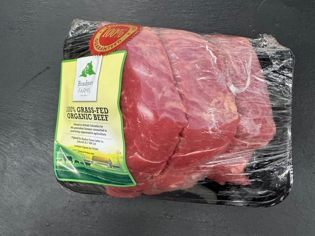 Packaged raw beef sirloin tip roast in a clear plastic-wrapped black tray with a green and yellow label reading '100% Grass Fed Organic Beef' and a red circular seal, placed on a dark gray surface.