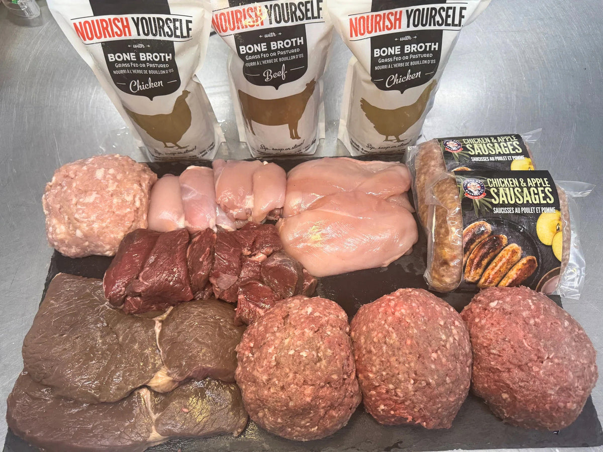A food preparation setup featuring raw meats and packaged products on a dark tray against a stainless steel background. Three white pouches of Nourish Yourself bone broth are lined up at the top, with raw chicken breasts, ground meat, beef chunks, and packaged chicken & apple sausages arranged below.