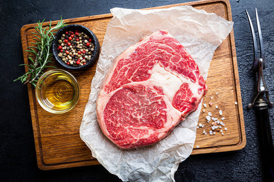 Ribeye steak thats raw, skits atop a cutting board, with herbs and spices, olive oil and seasoning