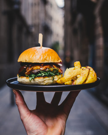 A hand holds up a plate with a burger and english style fries, while a street is barely visible in the background