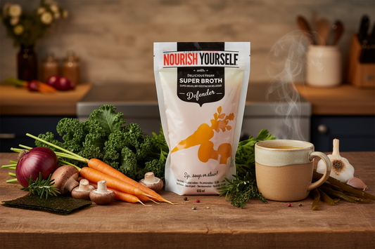 A packet of our vegan Defender Broth, alongside veggies and a mug filled with the hotr broth, on a wooden table in a kitchen.