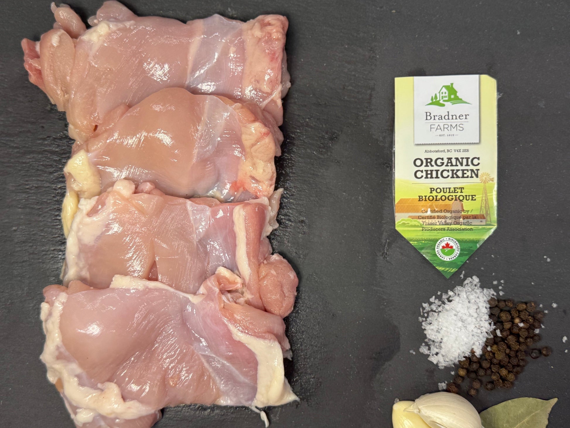 Four raw chicken thighs arranged on a dark slate surface, with a package of Dr. Brands Organic Paprika Seasoning, coarse salt, black peppercorns, a garlic bulb, and a bay leaf below.