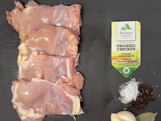 Four raw chicken thighs arranged on a dark slate surface, with a package of Dr. Brands Organic Paprika Seasoning, coarse salt, black peppercorns, a garlic bulb, and a bay leaf below.