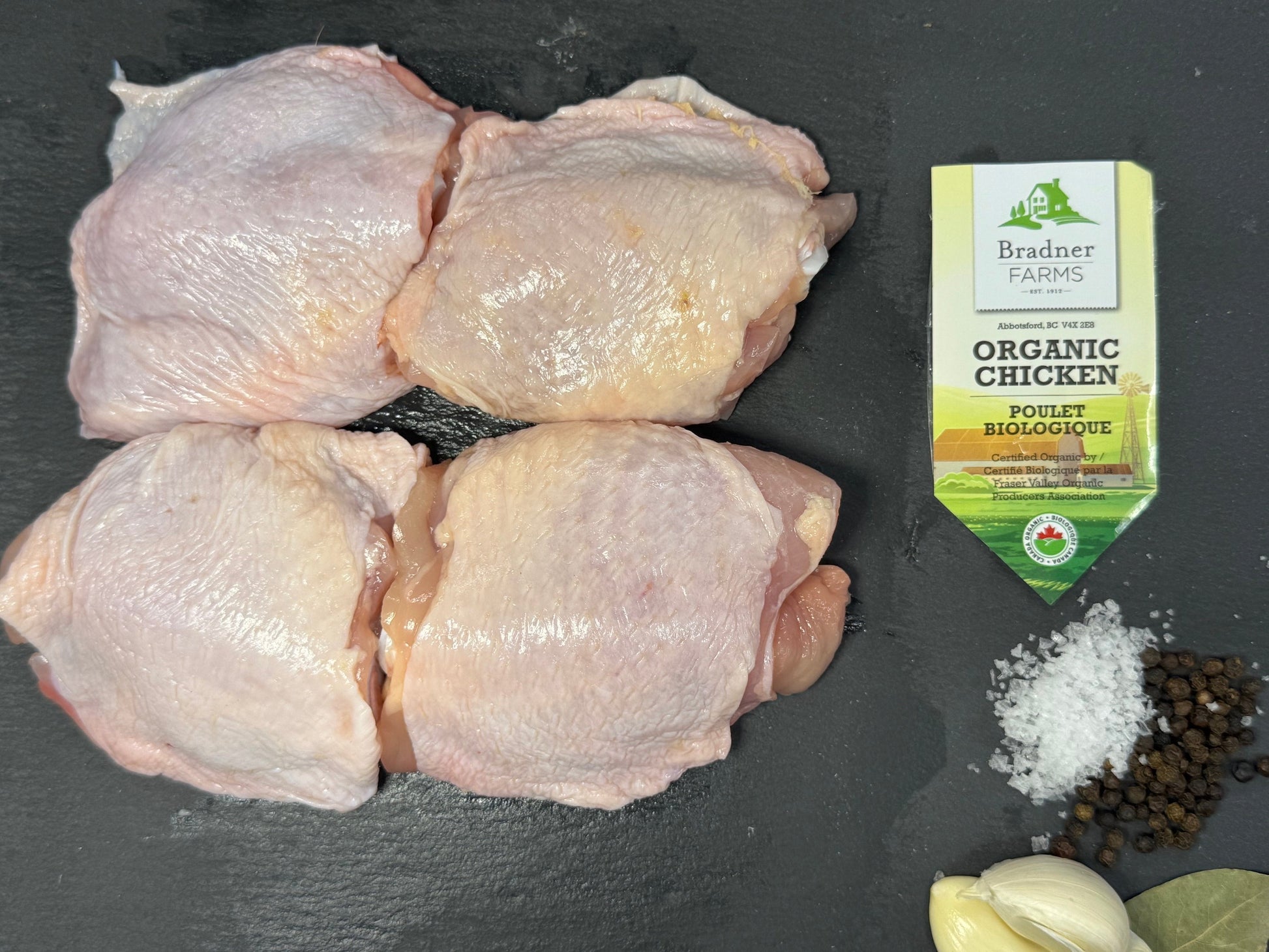 Four raw chicken thighs arranged on a dark slate surface, with a small package of Branded Farms Organic Chicken Seasoning, coarse salt, black peppercorns, and garlic cloves nearby.