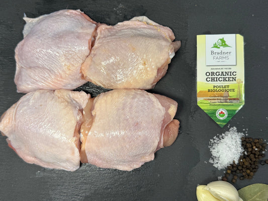 Four raw chicken thighs arranged on a dark slate surface, with a small package of Branded Farms Organic Chicken Seasoning, coarse salt, black peppercorns, and garlic cloves nearby.
