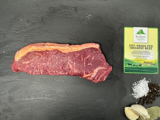 A raw beef steak is displayed on a dark slate surface next to garlic cloves, coarse sea salt, black peppercorns, and a package of Brisket Seasoning.