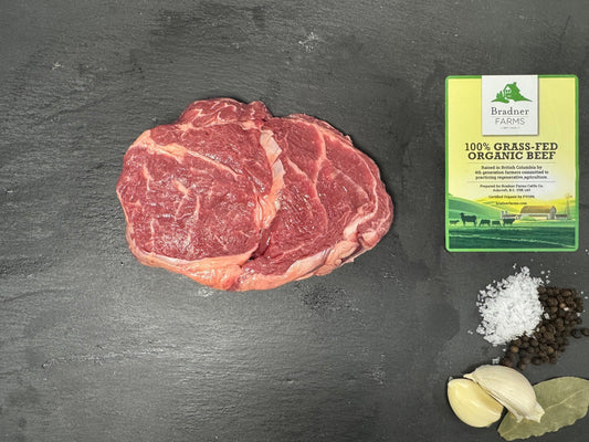 A raw cut of red beef steak on a dark slate surface, with a package of Brisket Ribs beef beside it and seasoning ingredients including salt, black pepper, garlic cloves, and bay leaves arranged around it.