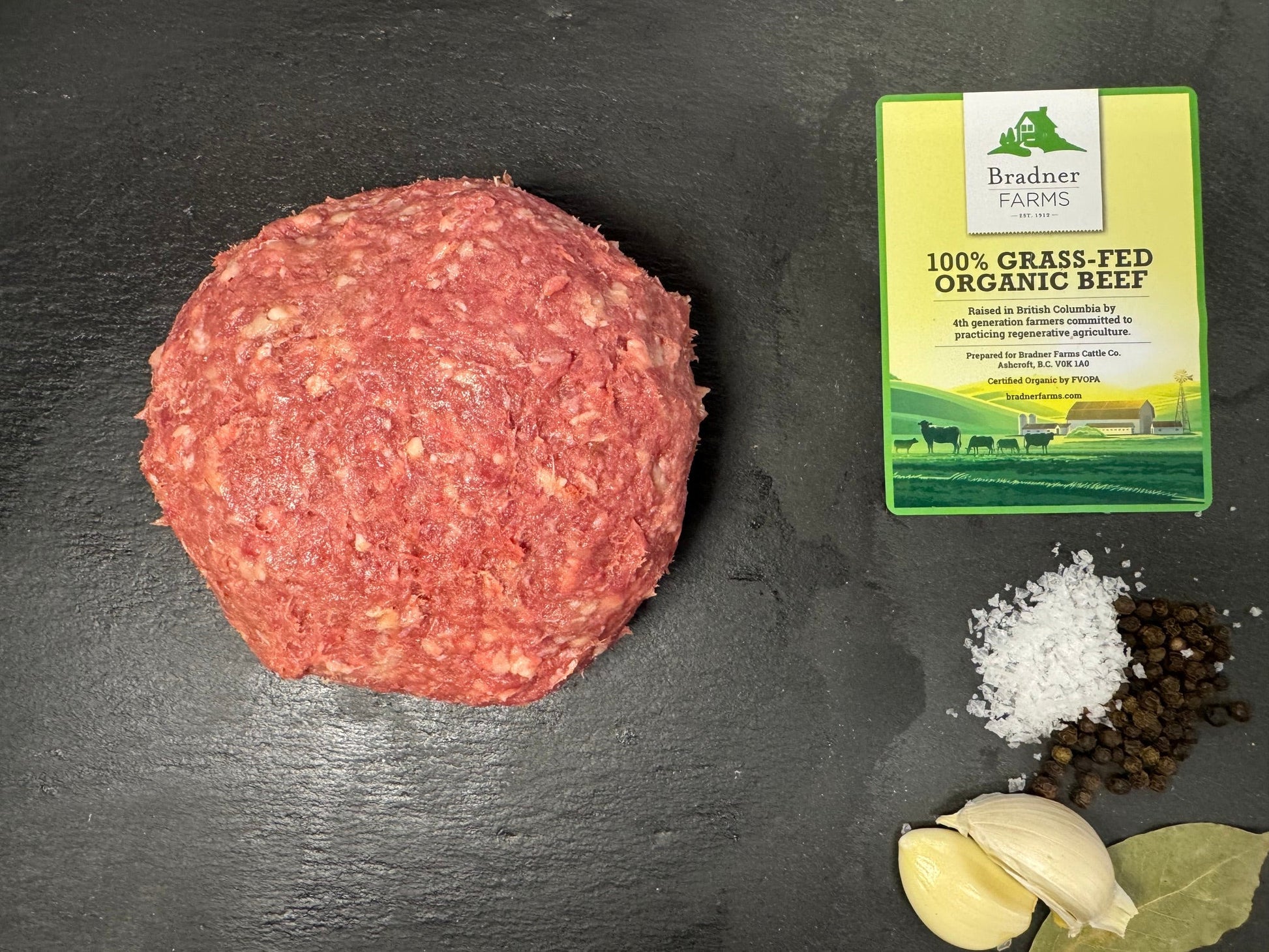 A raw ball of ground beef is displayed on a dark slate surface, with garlic cloves, coarse salt, black peppercorns, and a package of 100% grass-fed organic beef seasoning nearby.