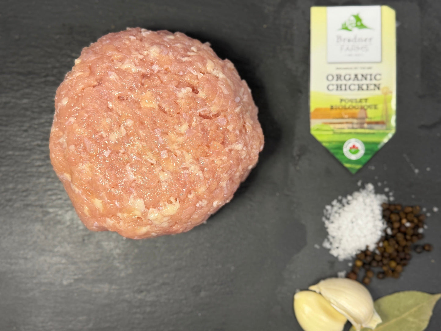 A raw round meatball sits on a dark slate surface, with garlic cloves, salt, peppercorns, and a small package of chicken broth nearby.