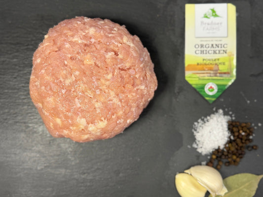 A raw round meatball sits on a dark slate surface, with garlic cloves, salt, peppercorns, and a small package of chicken broth nearby.