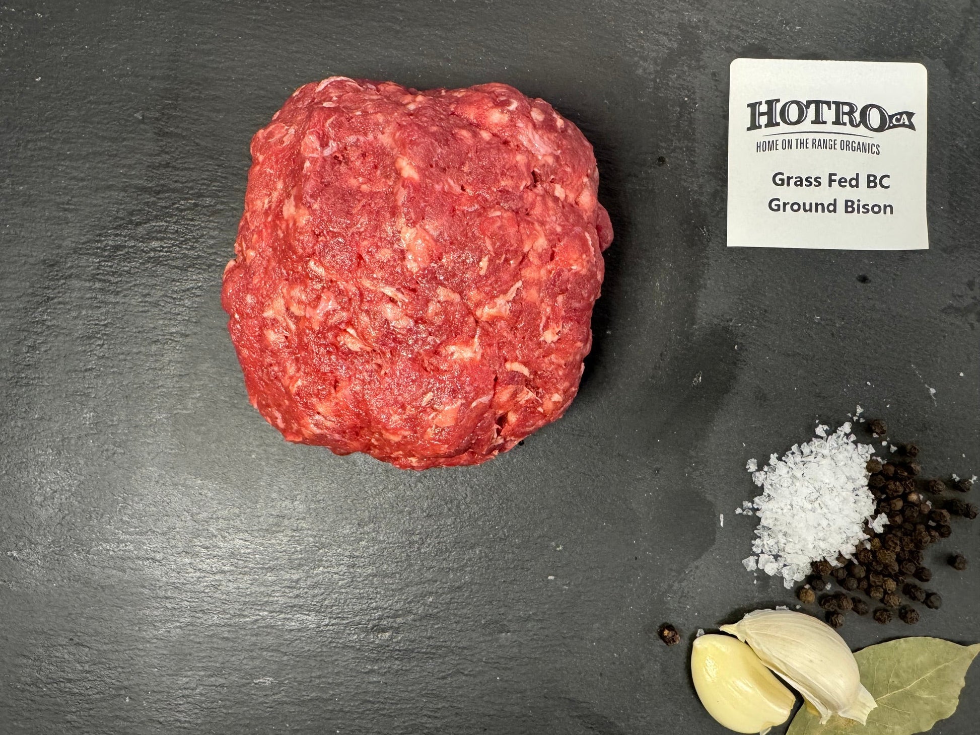 A raw round of ground beef, labeled HOTRO.ca Grass Fed BC Ground Bison, arranged on a dark slate background with garlic cloves, coarse salt, and peppercorns.