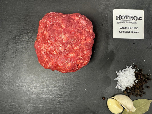 A raw round of ground beef, labeled HOTRO.ca Grass Fed BC Ground Bison, arranged on a dark slate background with garlic cloves, coarse salt, and peppercorns.