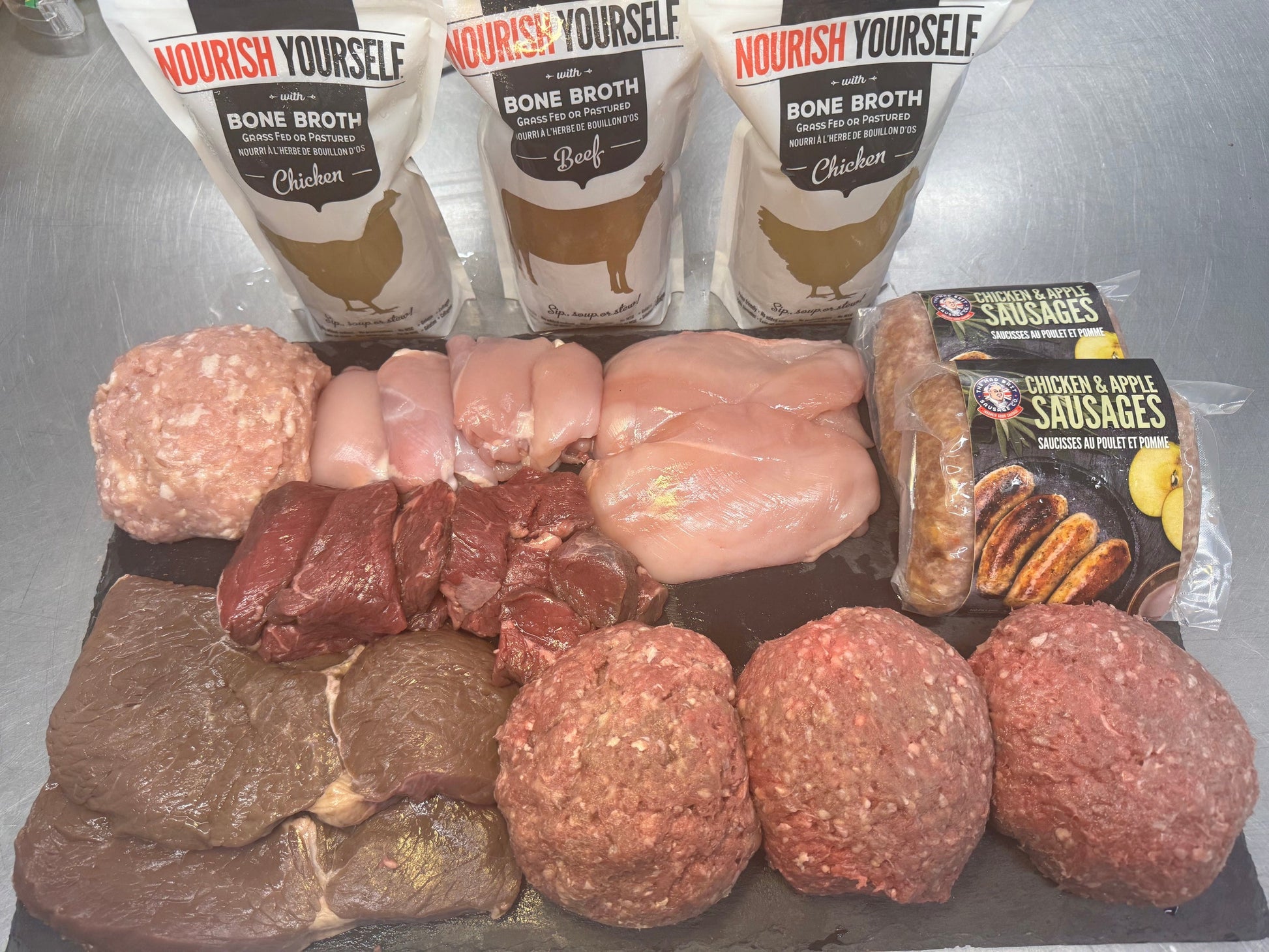 An assortment of grass fed beef and organic chicken on a slate board, with Nourish Yourself Bone Broth (beef and chicken), plus Mad Brit Chicken and Apple Sausages