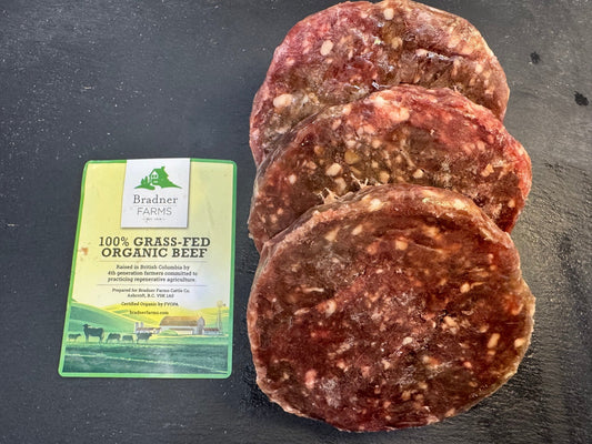 Three raw beef burger patties with visible fat marbling are arranged on a dark surface next to a green and white package labeled '100% Grass Fed Organic Beef' by Bradner Farms.