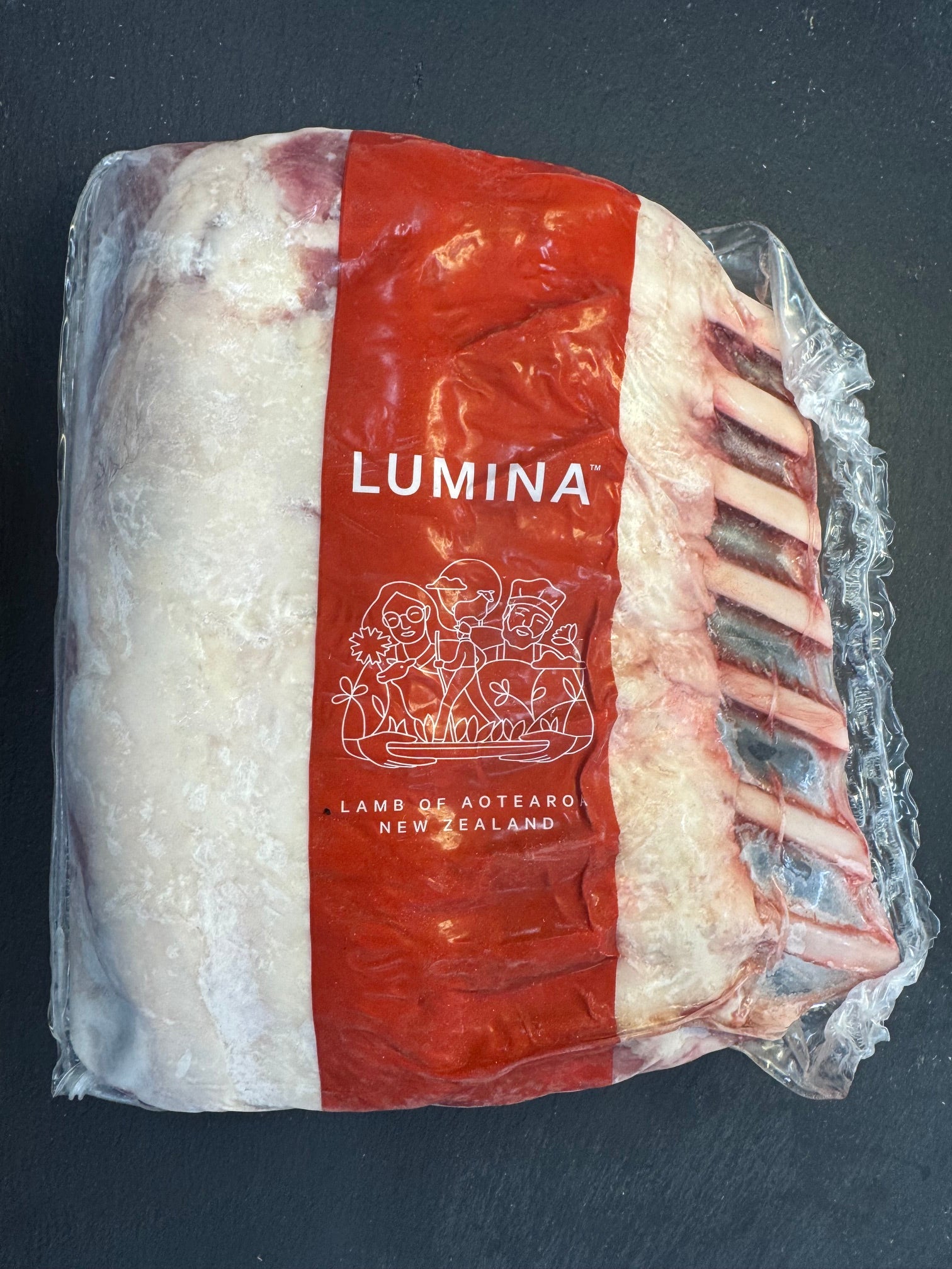 A vacuum-sealed package of Lumina Lamb from New Zealand meat, showing the white marbled fat and red meat layers with visible bones on the side, set against a dark slate background.