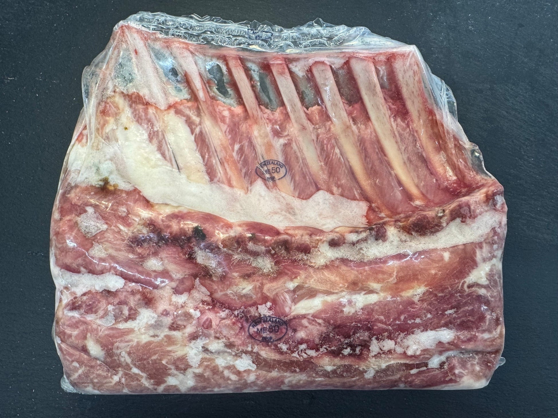 A packaged raw cut of rack of lamb with visible ribs and marbled fat, sealed in clear plastic on a dark surface.