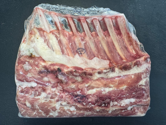 A packaged raw cut of rack of lamb with visible ribs and marbled fat, sealed in clear plastic on a dark surface.