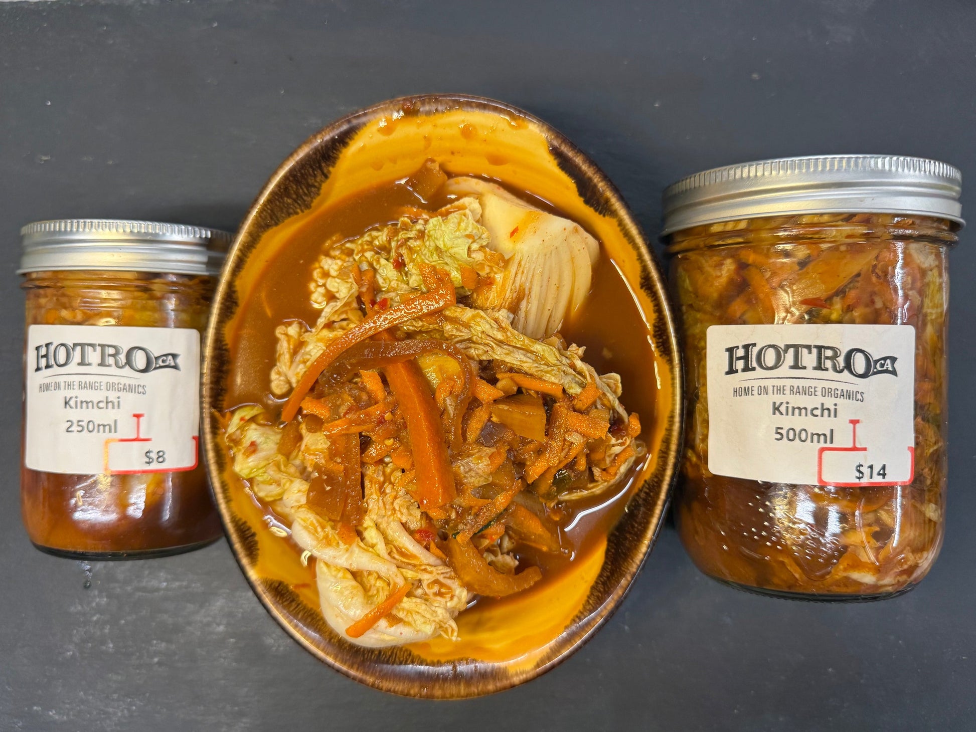 Three glass jars of Hotoga Kimchi with silver lids are arranged around a ceramic bowl filled with the prepared kimchi dish, set on a dark gray surface.