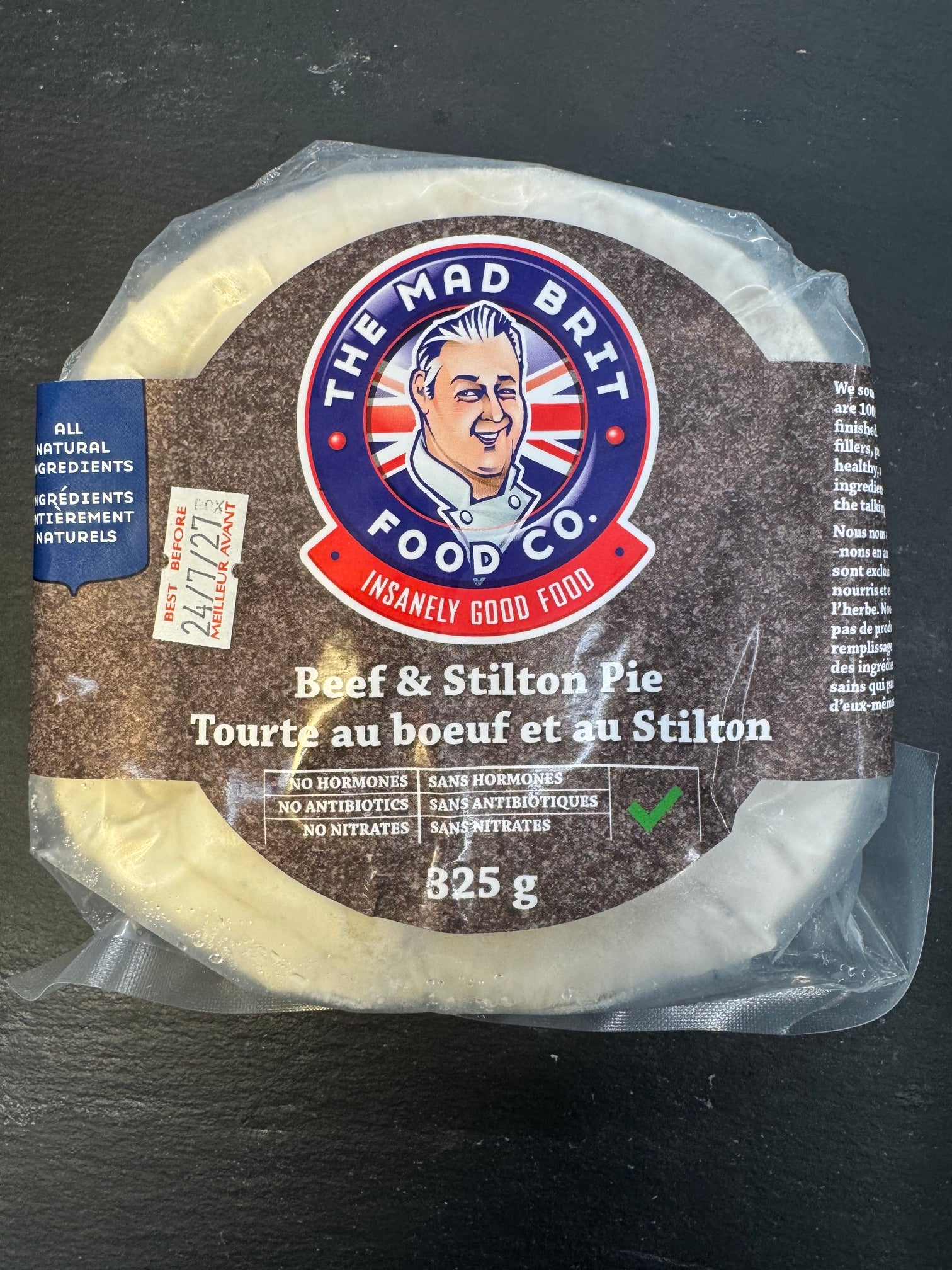 A packaged round cheese product labeled 'The Mad Brit Food Co.' and 'Beef & Stilton Pie' on a dark textured surface.