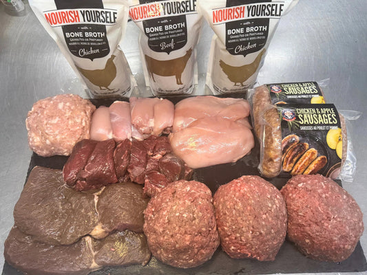 A food preparation setup featuring raw meats and packaged products on a dark tray against a stainless steel background. Three white pouches of Nourish Yourself bone broth are lined up at the top, with raw chicken breasts, ground meat, beef chunks, and packaged chicken & apple sausages arranged below.